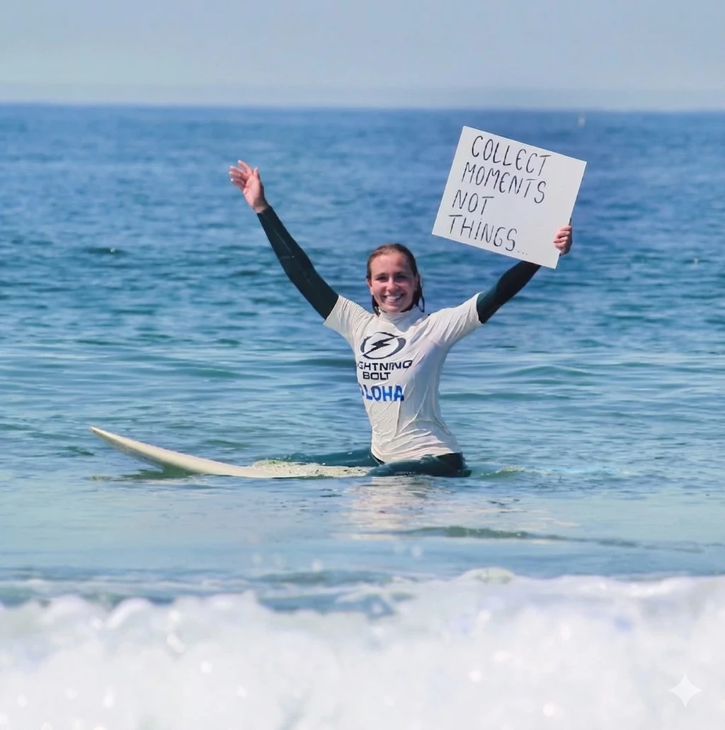 Surfista sentada na sua prancha no mar com os braços erguidos a segurar um cartaz que diz 'collect moments not things', a sorrir após uma onda.